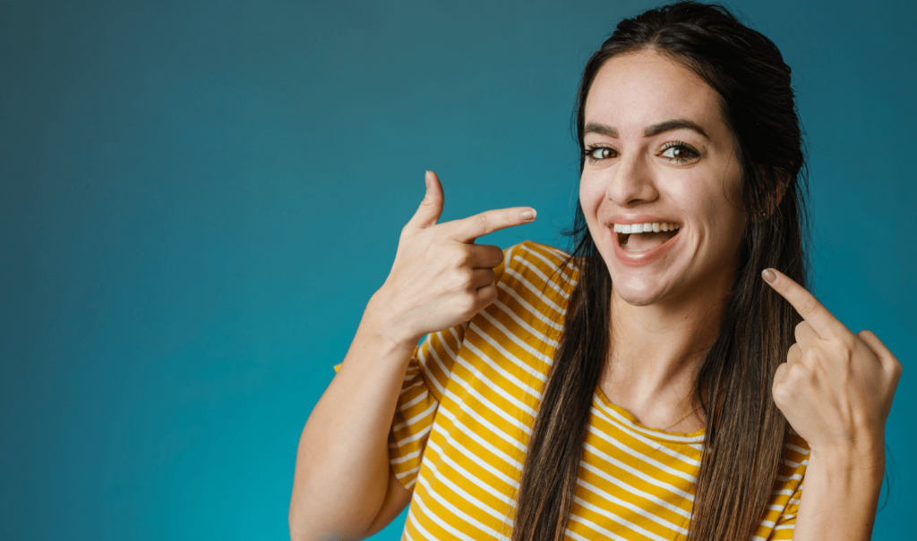 A girl standing agaisnt a blue background, and pointing at her smile. She has long hair and is wearing a yellow shirt.
